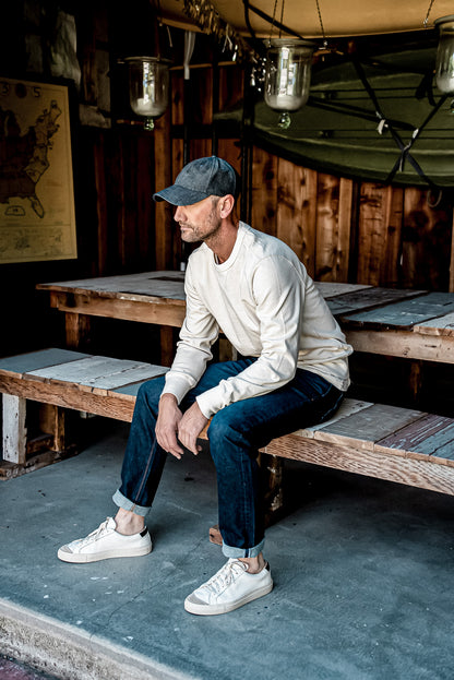Man in cuffed dark ocean wash American made denim jeans, cream sweatshirt and cap, seated on a wooden bench.