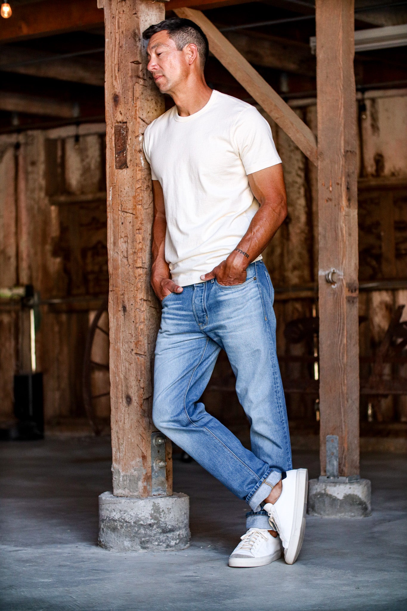 Man in salt wash Sam standard fit jeans and cream tee, leaning against a barn beam.