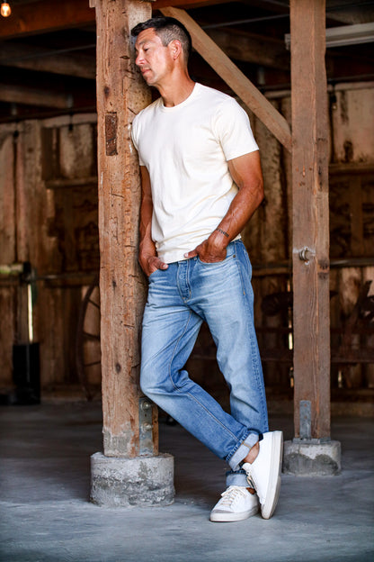 Man in salt wash Sam standard fit jeans and cream tee, leaning against a barn beam.