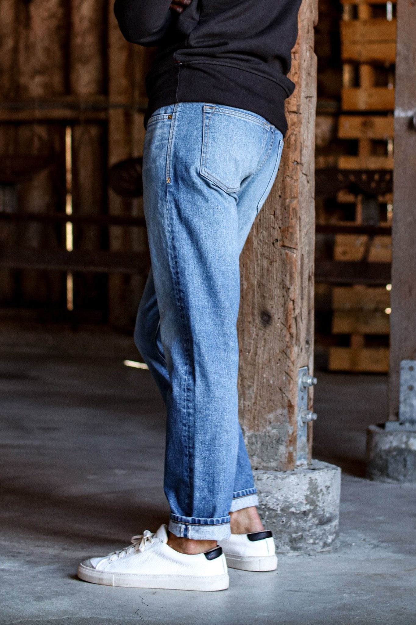 Side view of salt wash American made denim jeans with cuffed hem, worn with white sneakers in a rustic warehouse.