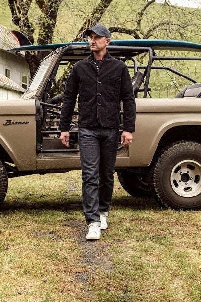 Man in black denim chore blazer walking past a vintage Bronco with surfboard.