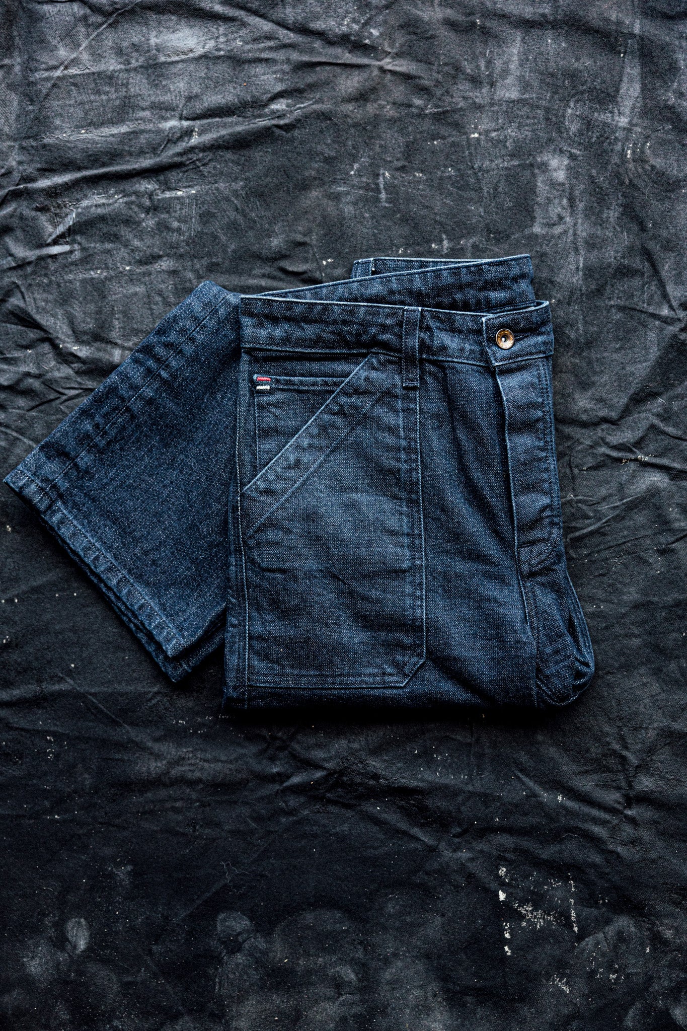 Women’s premium denim jeans folded neatly on a dark textured surface, showcasing rich blue fabric, structured seams, and front utility pocket detailing