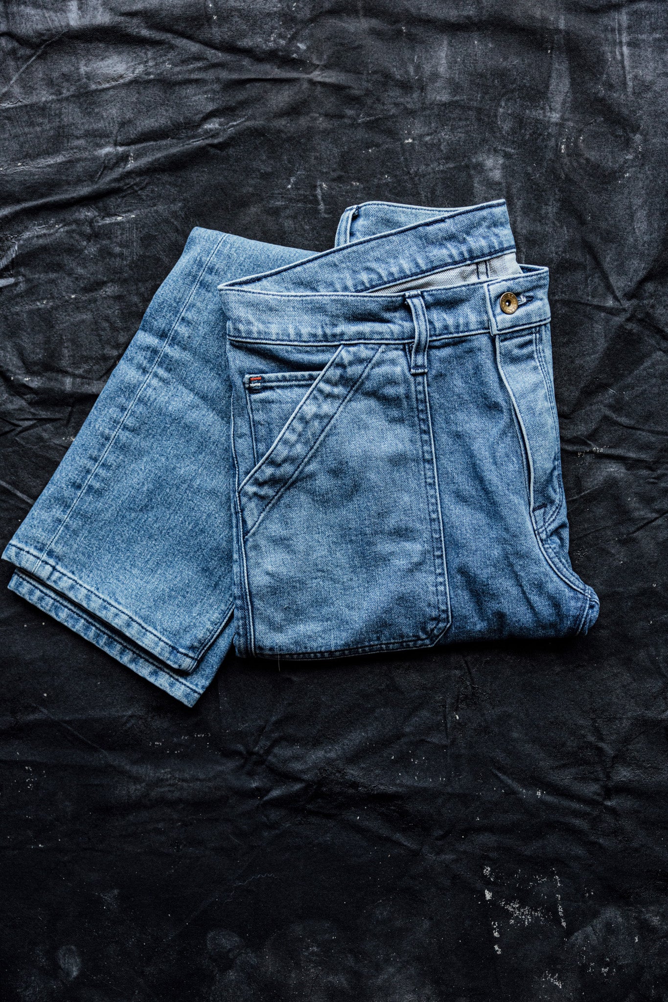 Women’s premium denim jeans folded on a dark textured surface, showcasing light blue salt wash fabric, clean seams, and structured utility pocket detailing