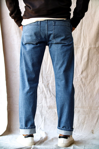 Raw denim jeans with a straight-leg fit, back patch pockets, and cuffed hems, styled with white sneakers and a black sweatshirt.