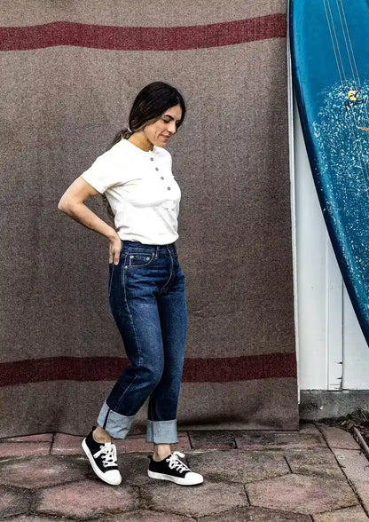 Model in high-rise selvedge denim jeans with cuffed hems, white henley tee, and black sneakers beside a surfboard.