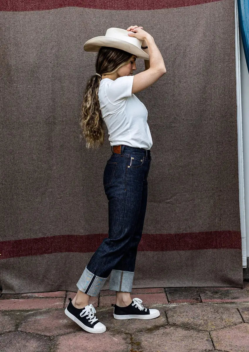 Side view of high-rise raw selvedge denim jeans with cuffed hem, worn with a white tee, cowboy hat, and black sneakers.