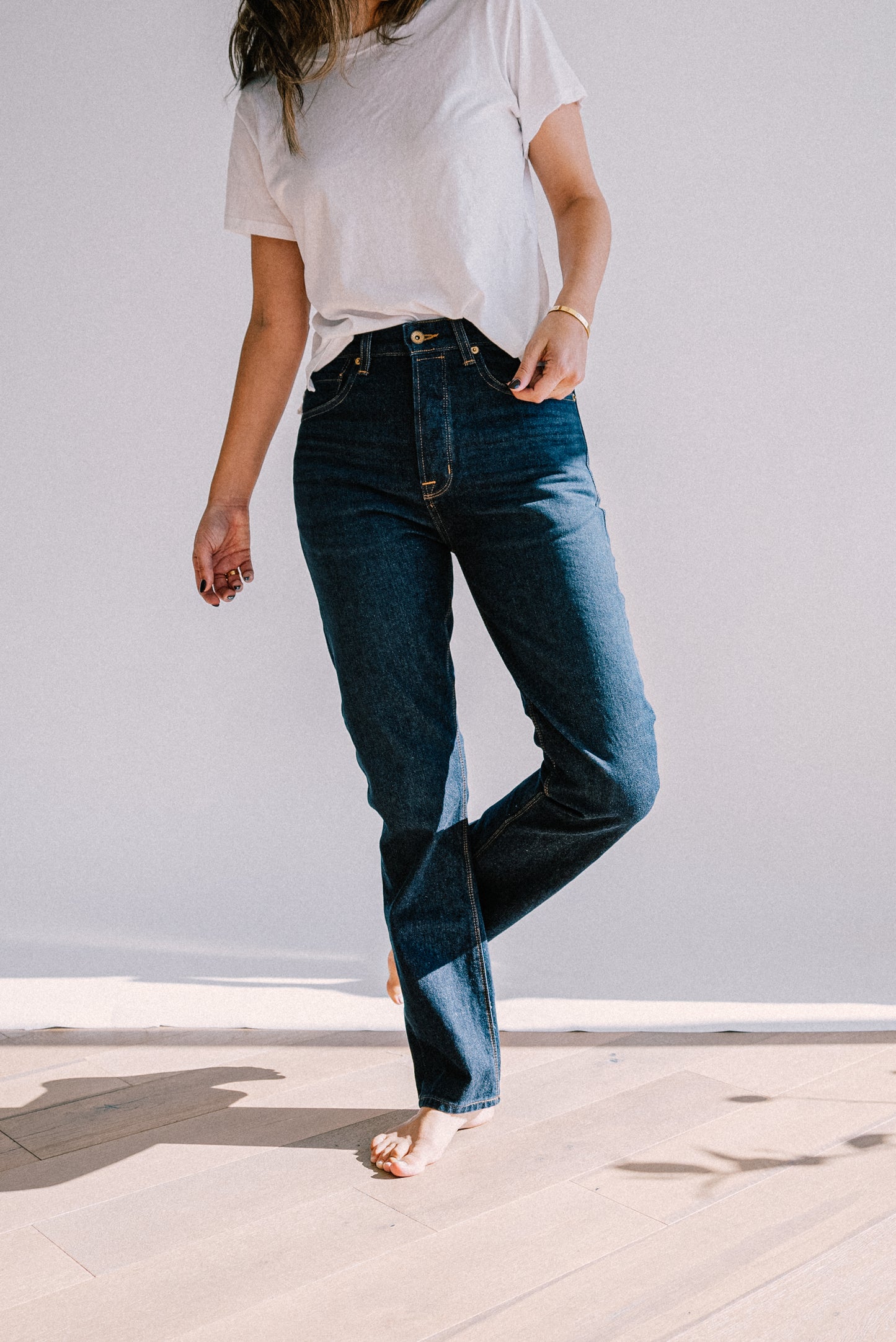 Model in a white tee and dark ocean wash high-rise straight-leg American made jeans, barefoot on light wood floor.