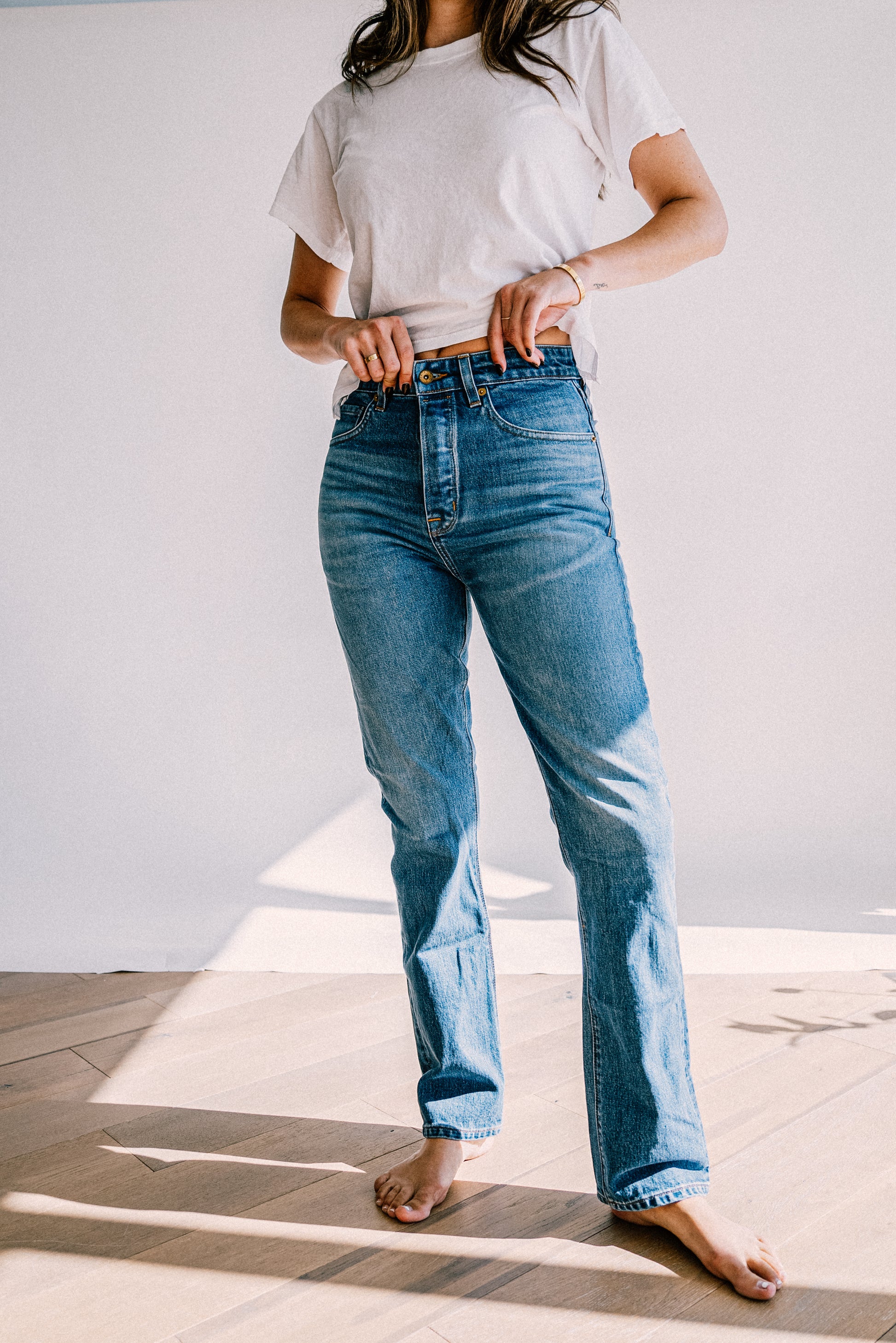 Model in a white tee and salt wash high-rise straight leg American made jeans, barefoot on a sunlit wood floor.