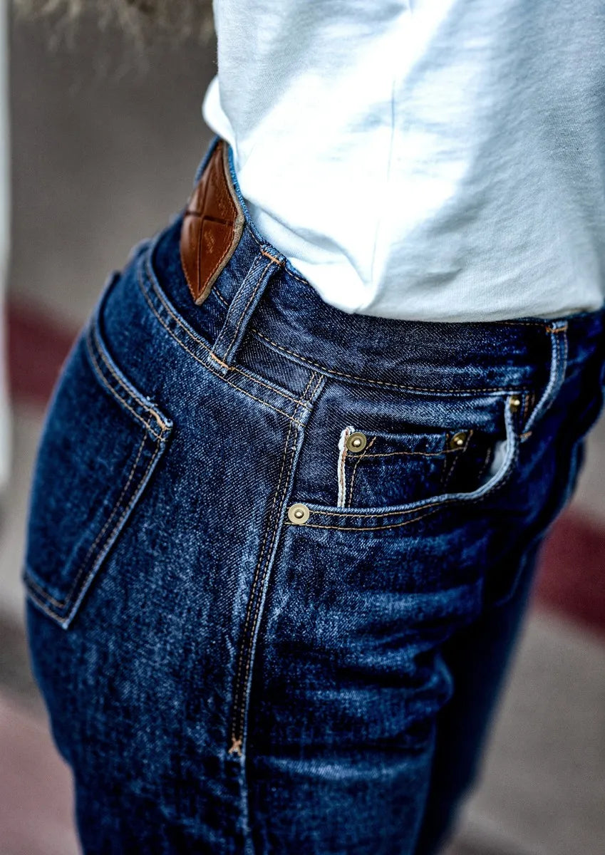 Close-up of high-rise selvedge denim jeans showing front pocket, brass rivets, and leather patch at the waistband.