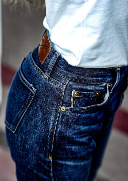 Close-up of high-rise selvedge denim jeans showing front pocket, brass rivets, and leather patch at the waistband.