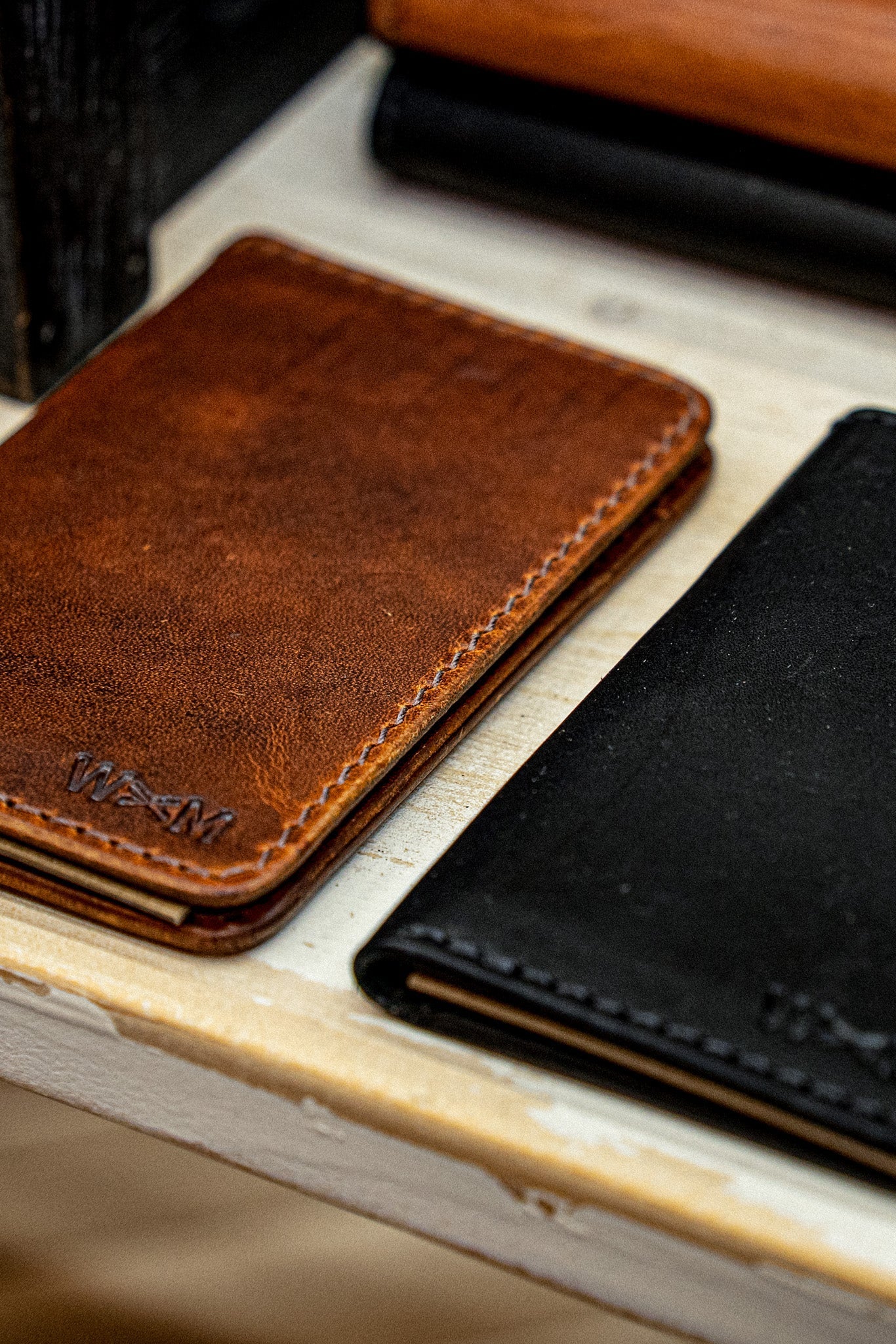 Tan Horween leather small wallet with embossed logo beside a black wallet on wood.