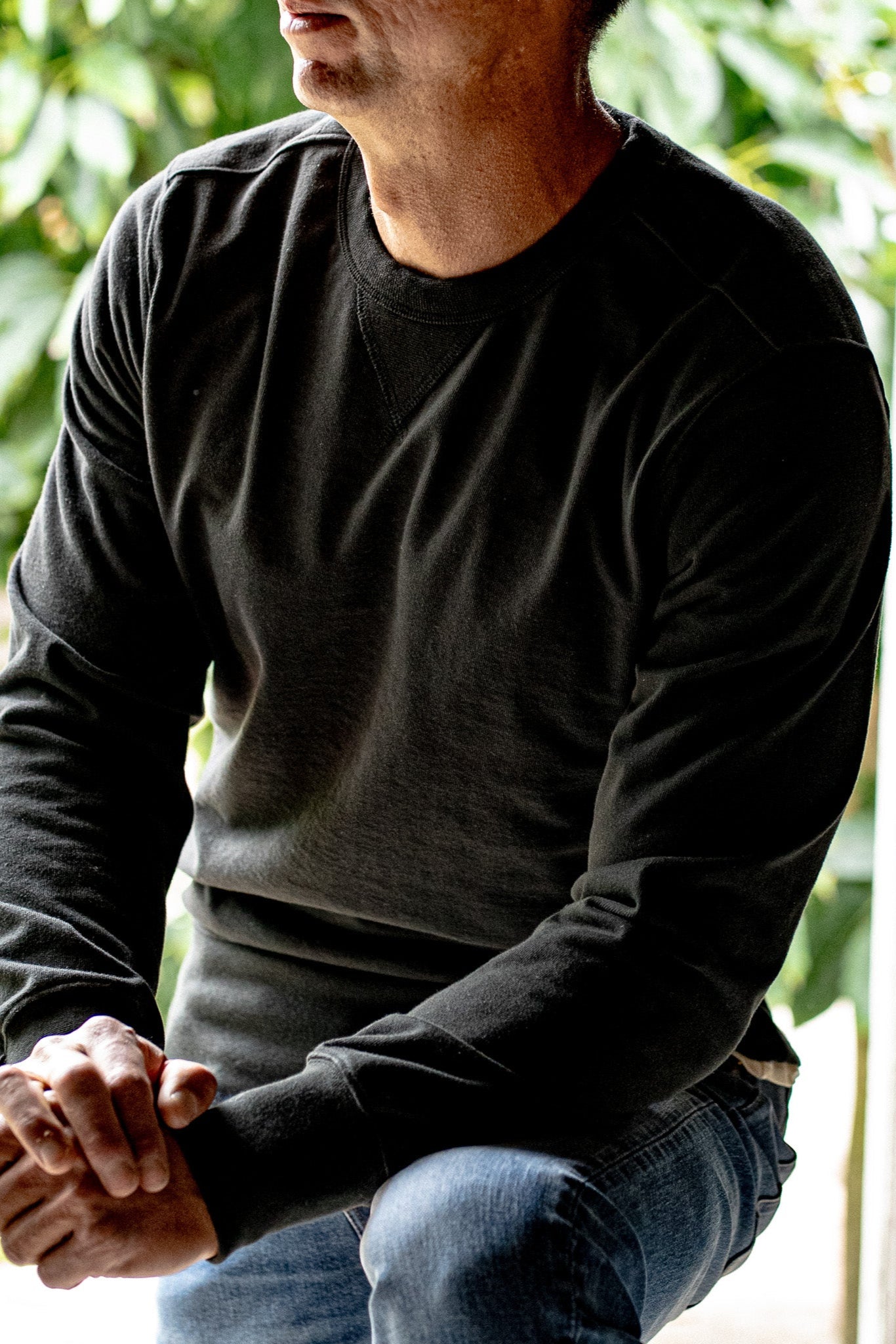 Man wearing a basalt black Stinson rugby jersey pullover with blue denim jeans, seated outdoors.