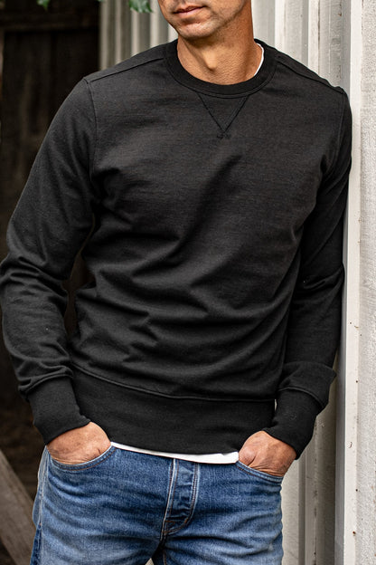 Men's Stinson rugby jersey pullover in basalt black, worn with blue denim jeans against a weathered wood wall.