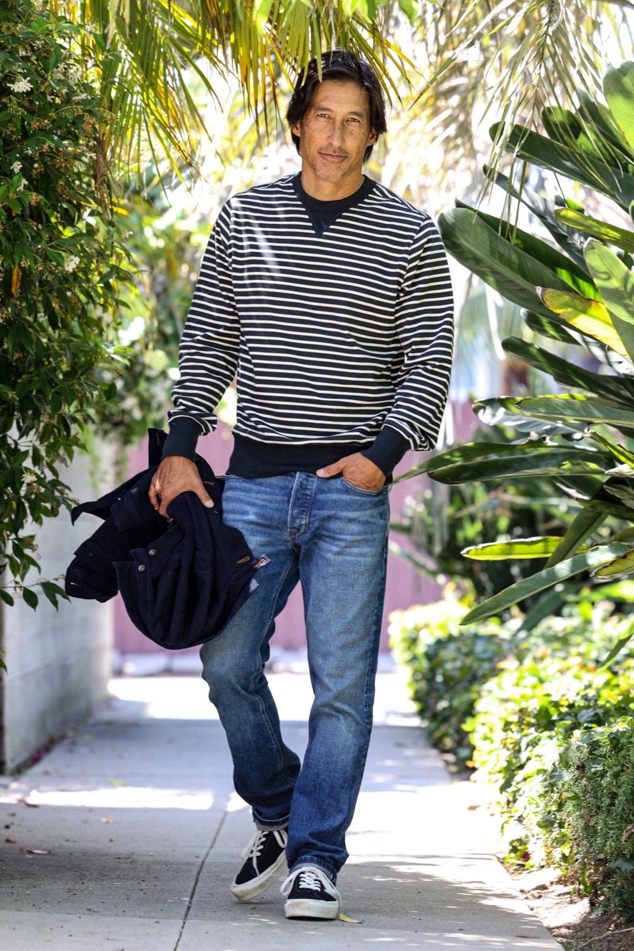 Man in navy striped Stinson rugby jersey pullover walking a sunlit palm-lined sidewalk.