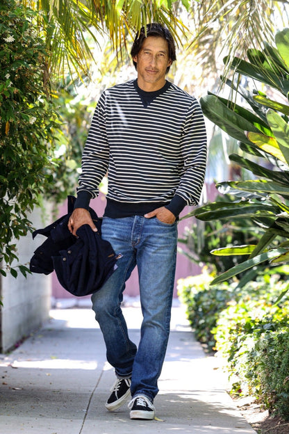 Man in navy striped Stinson rugby jersey pullover walking a sunlit palm-lined sidewalk.