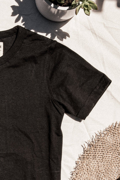 Basalt black heavyweight cotton tee laid flat beside a succulent and woven straw hat in sunlight.