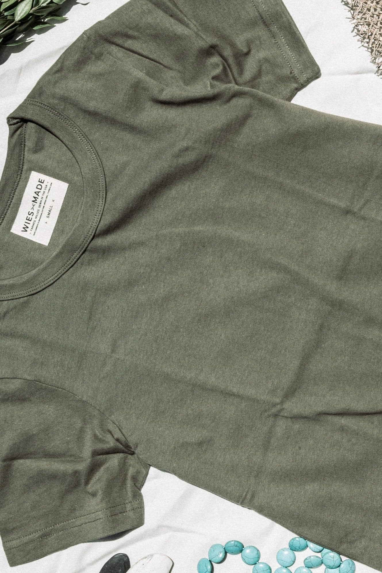 Olive green heavyweight cotton tee laid flat with WiesMade neck label, beside greenery and turquoise stones.