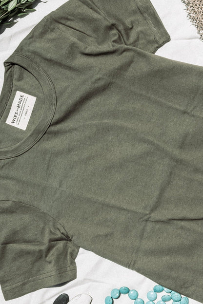 Olive green heavyweight cotton tee laid flat with WiesMade neck label, beside greenery and turquoise stones.
