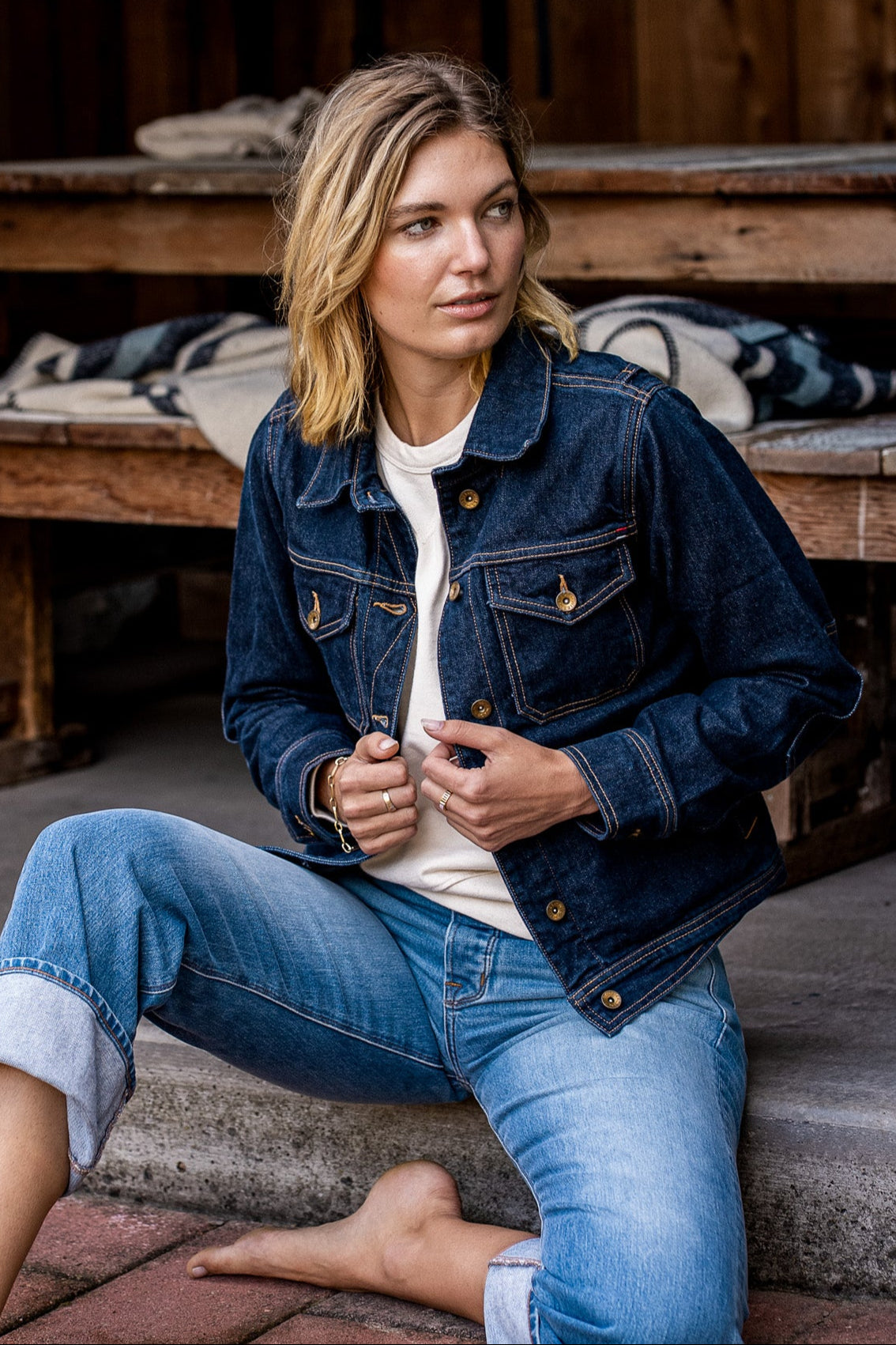 Woman wearing the Artcher broken twill denim jacket in ocean blue with brass buttons, seated on a porch.