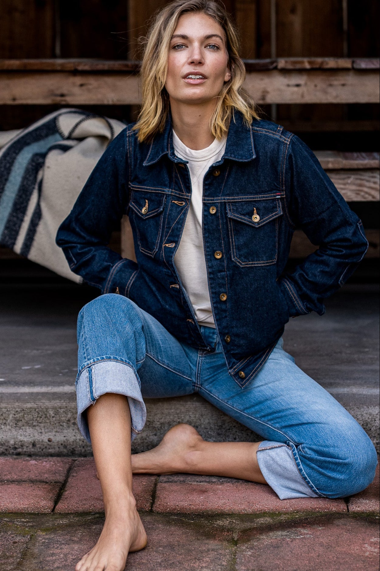 Woman in dark ocean-wash broken twill denim jacket and cuffed jeans seated on brick steps.