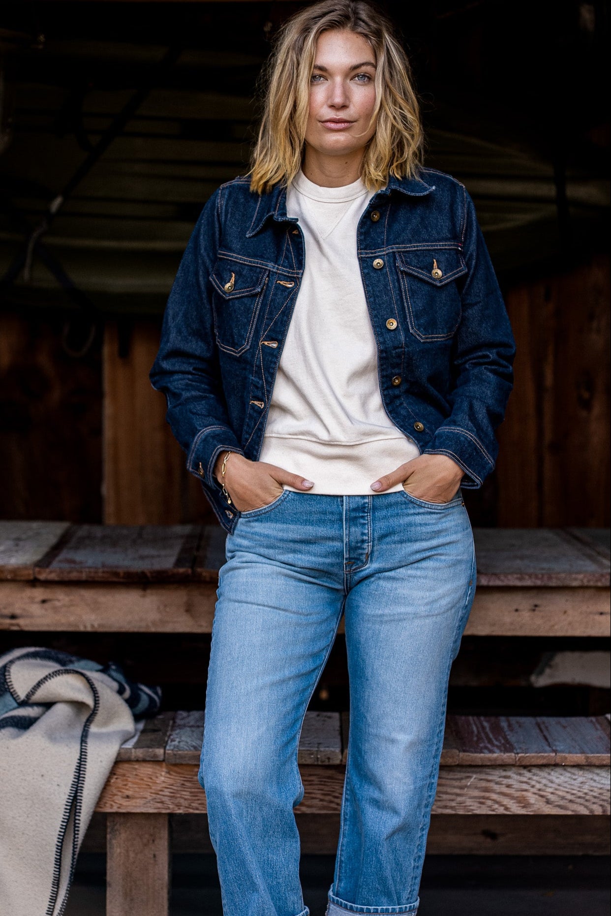 Woman wearing the ocean-wash broken twill denim jacket over a cream sweatshirt with light blue jeans on a rustic porch.