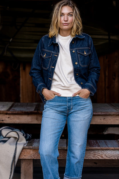 Woman wearing the ocean-wash broken twill denim jacket over a cream sweatshirt with light blue jeans on a rustic porch.