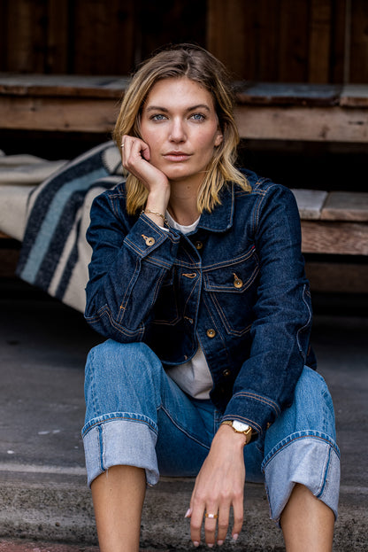 Woman seated on wooden steps wearing the Artcher broken twill denim jacket in Ocean with brass buttons and contrast.
