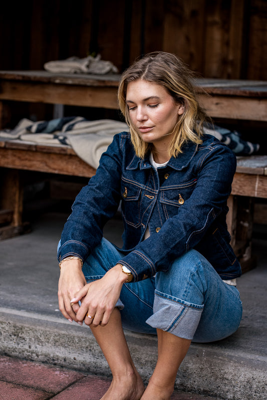 Model seated on a step wearing the Artcher broken twill denim jacket in ocean with contrast stitching and brass buttons.