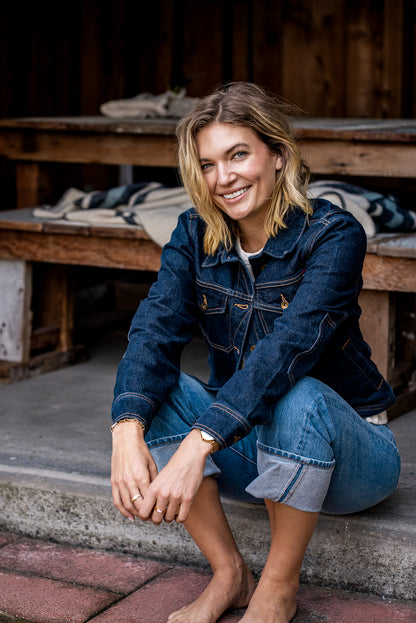 Smiling woman crouched on a porch wearing the ocean-blue broken twill denim jacket with cuffed jeans.
