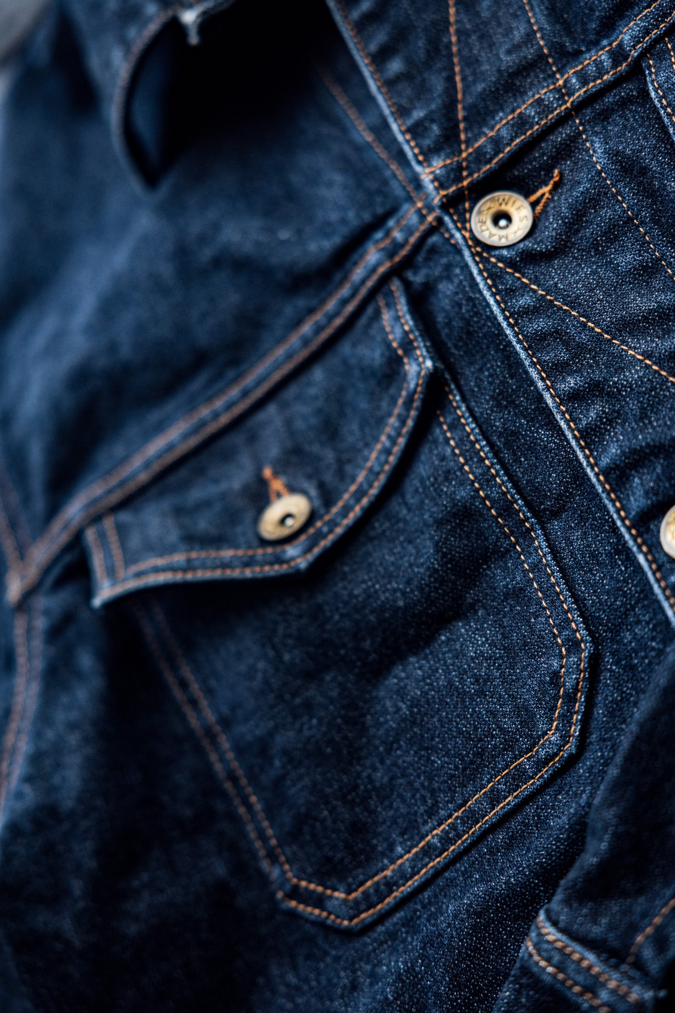 Close-up of the Artcher broken twill denim jacket showing contrast orange stitching and brass chest pocket button.
