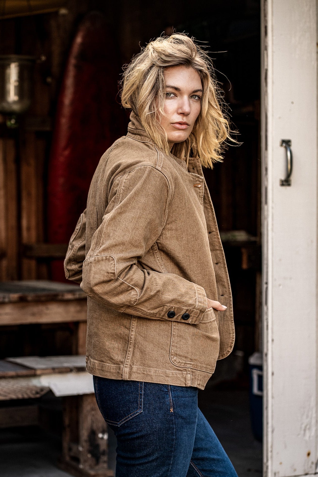 Woman in wheat denim chore blazer with buttoned cuff, standing in a rustic doorway.