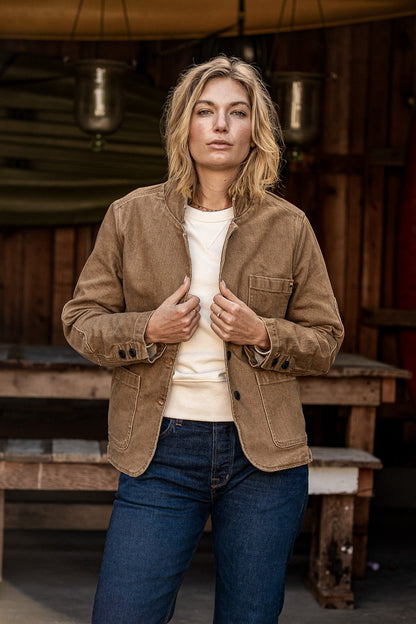 Woman wearing wheat denim Scout Blazer over cream tee and jeans at a rustic porch.