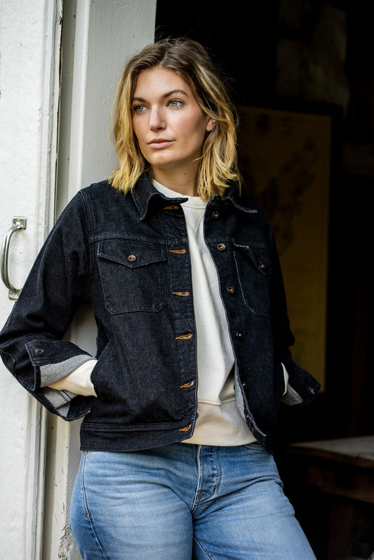 Woman leaning in a doorway wearing the black broken twill denim Artcher Jacket with tobacco keyholes over a cream sweatshirt.