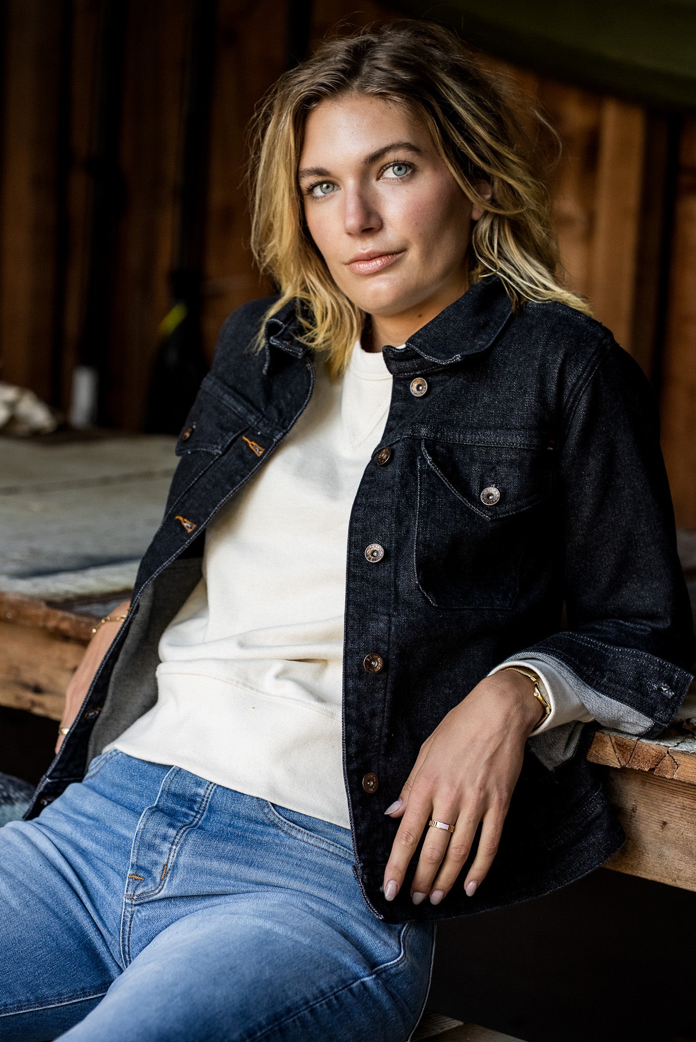 Woman in a black broken twill denim jacket over a cream sweatshirt, seated on a wooden bench.