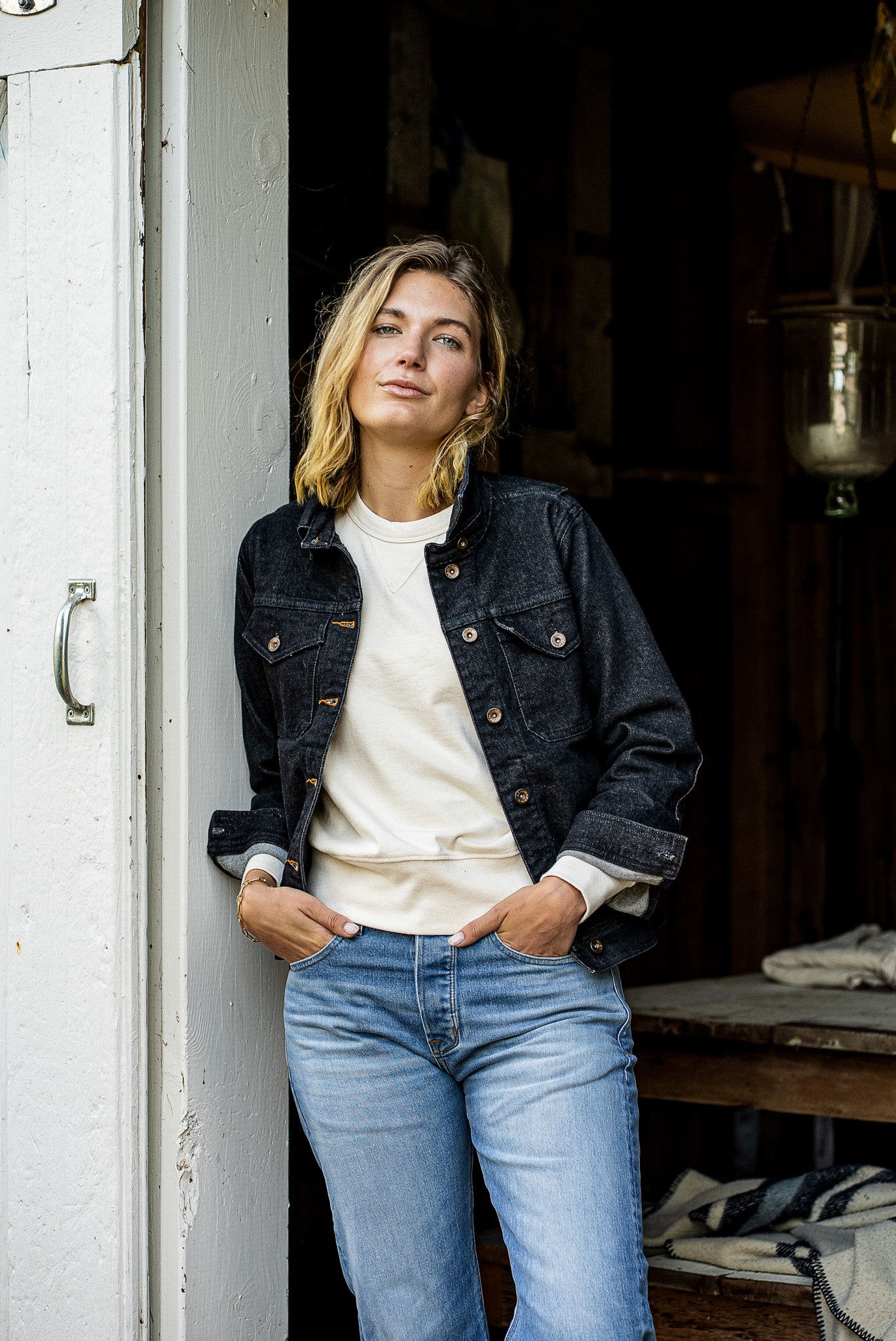 Woman leaning in a barn doorway wearing a black broken twill denim jacket over a cream sweatshirt and light blue jeans.