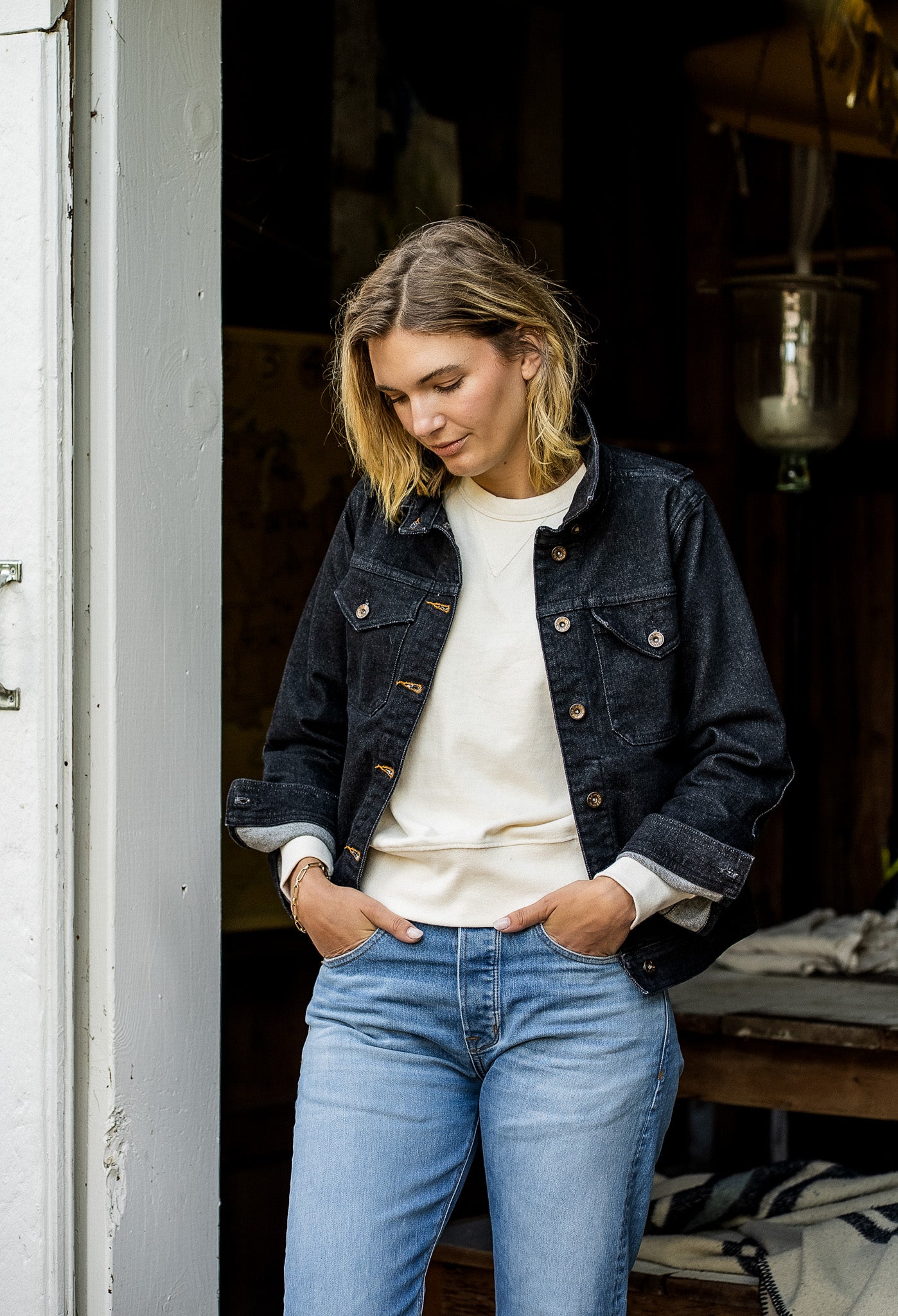 Woman wearing a black broken twill denim jacket over a cream sweatshirt with light wash jeans in a doorway.