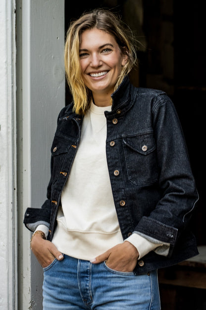 Smiling woman wearing a black broken twill denim jacket over a cream sweatshirt with blue jeans.