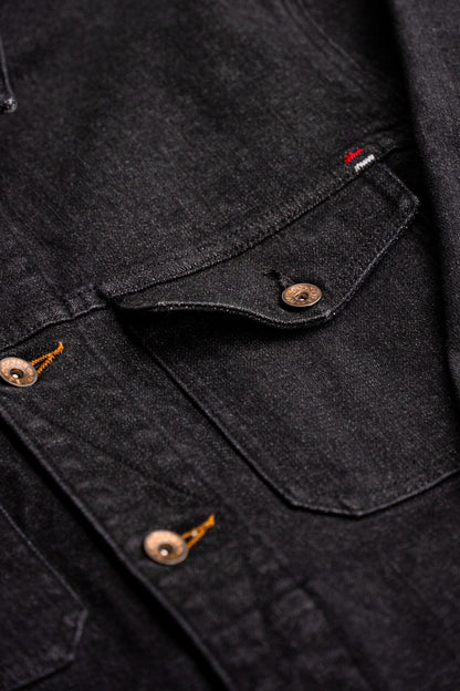 Close-up of black broken twill denim jacket showing chest pocket, brass buttons, and tobacco keyhole stitching.