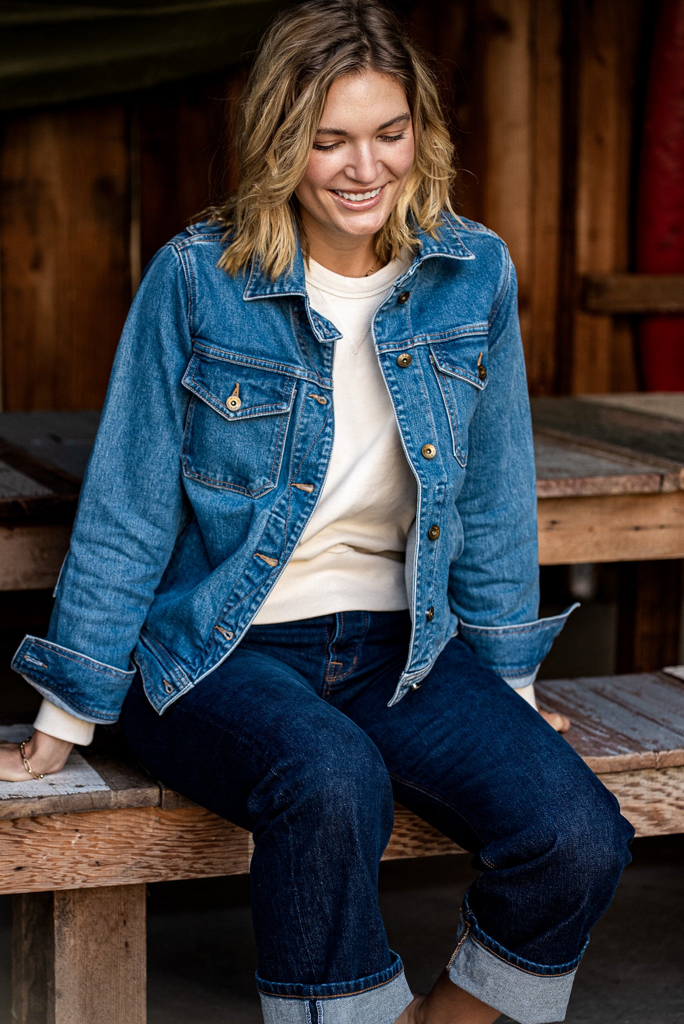 Woman in a light-wash broken twill denim jacket and cuffed dark jeans, seated on a wooden bench.