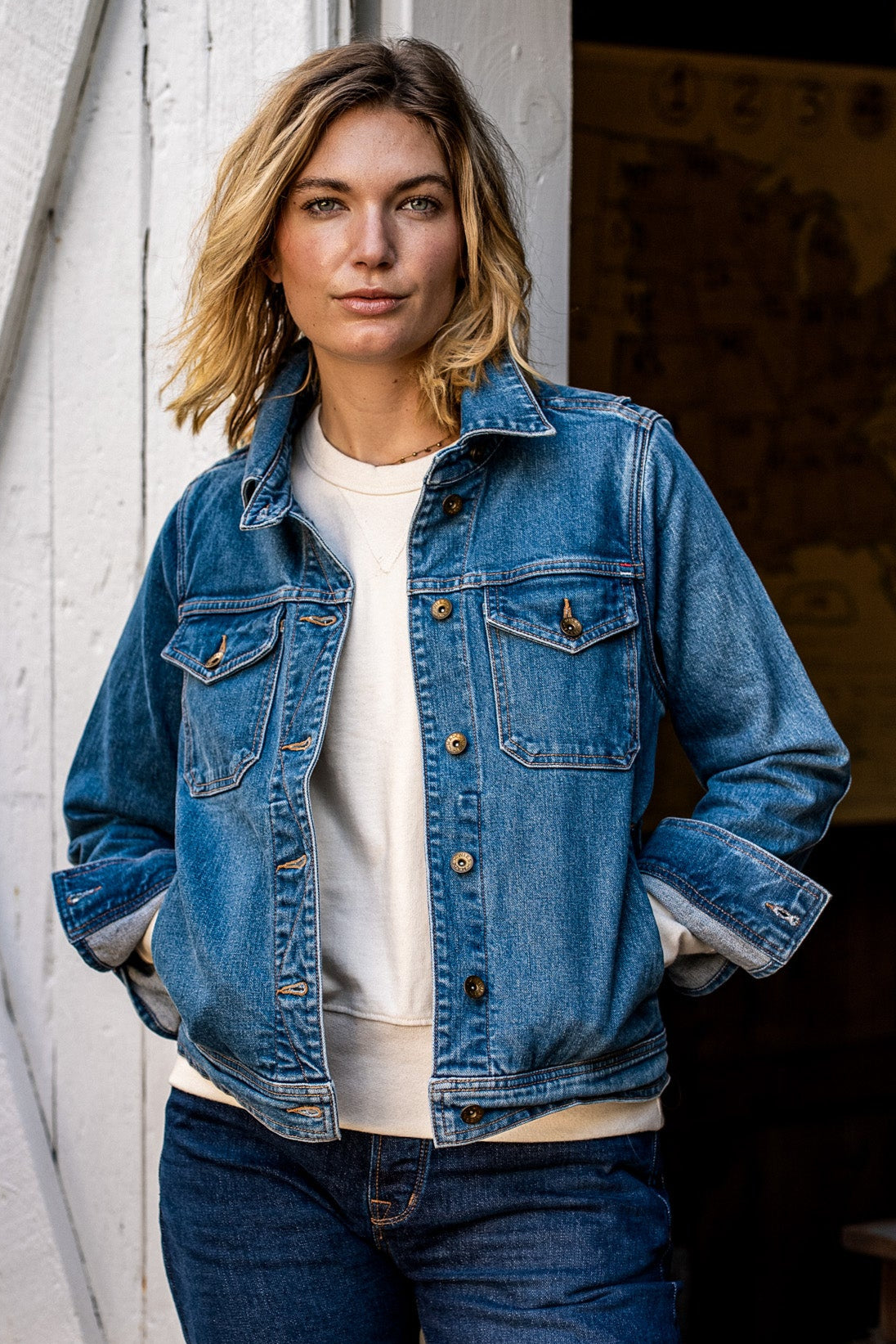Woman wearing the Artcher broken twill denim jacket in salt wash over a cream tee, standing by a weathered white barn door.