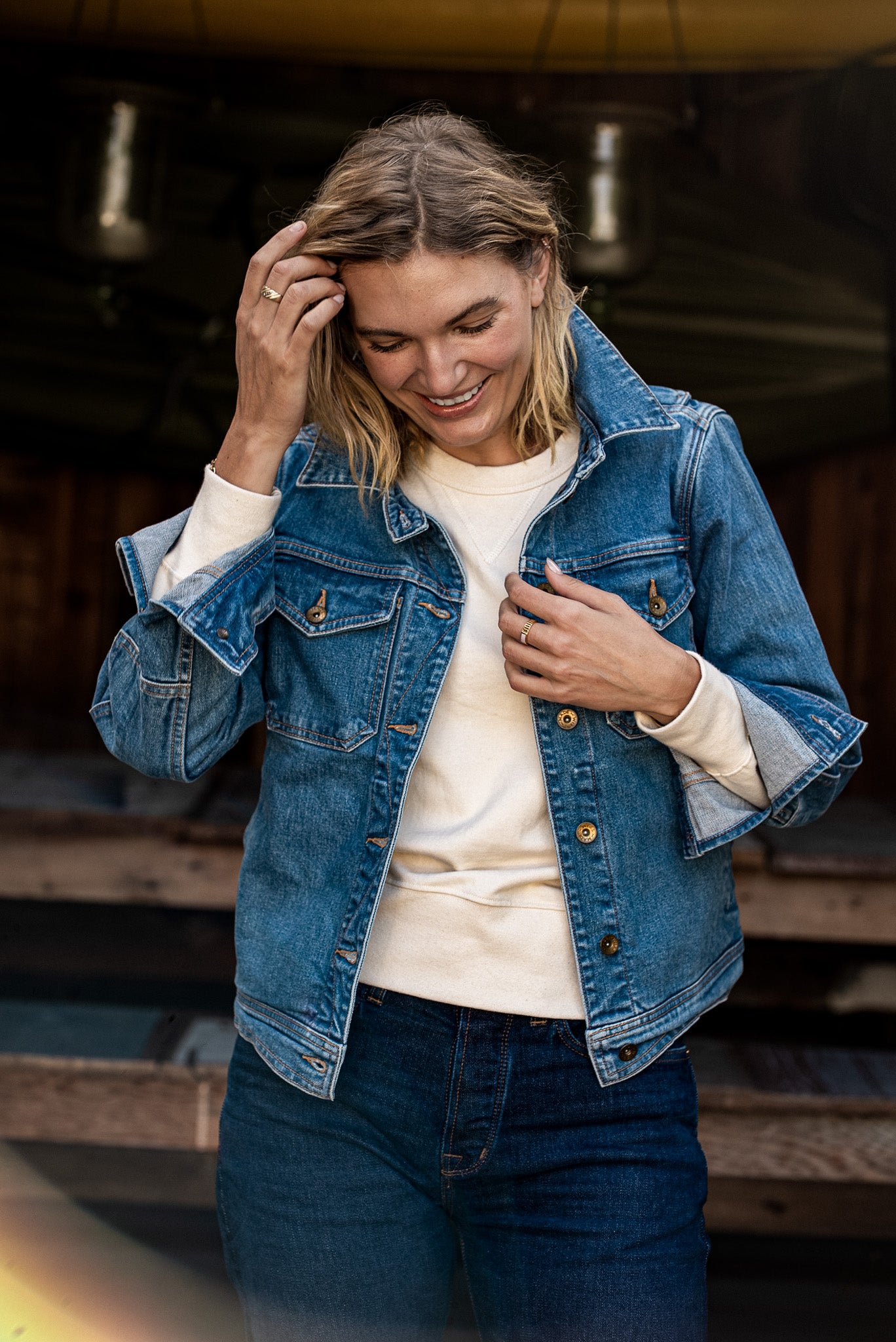 Woman wearing an open broken twill denim jacket over a cream sweatshirt and dark jeans.