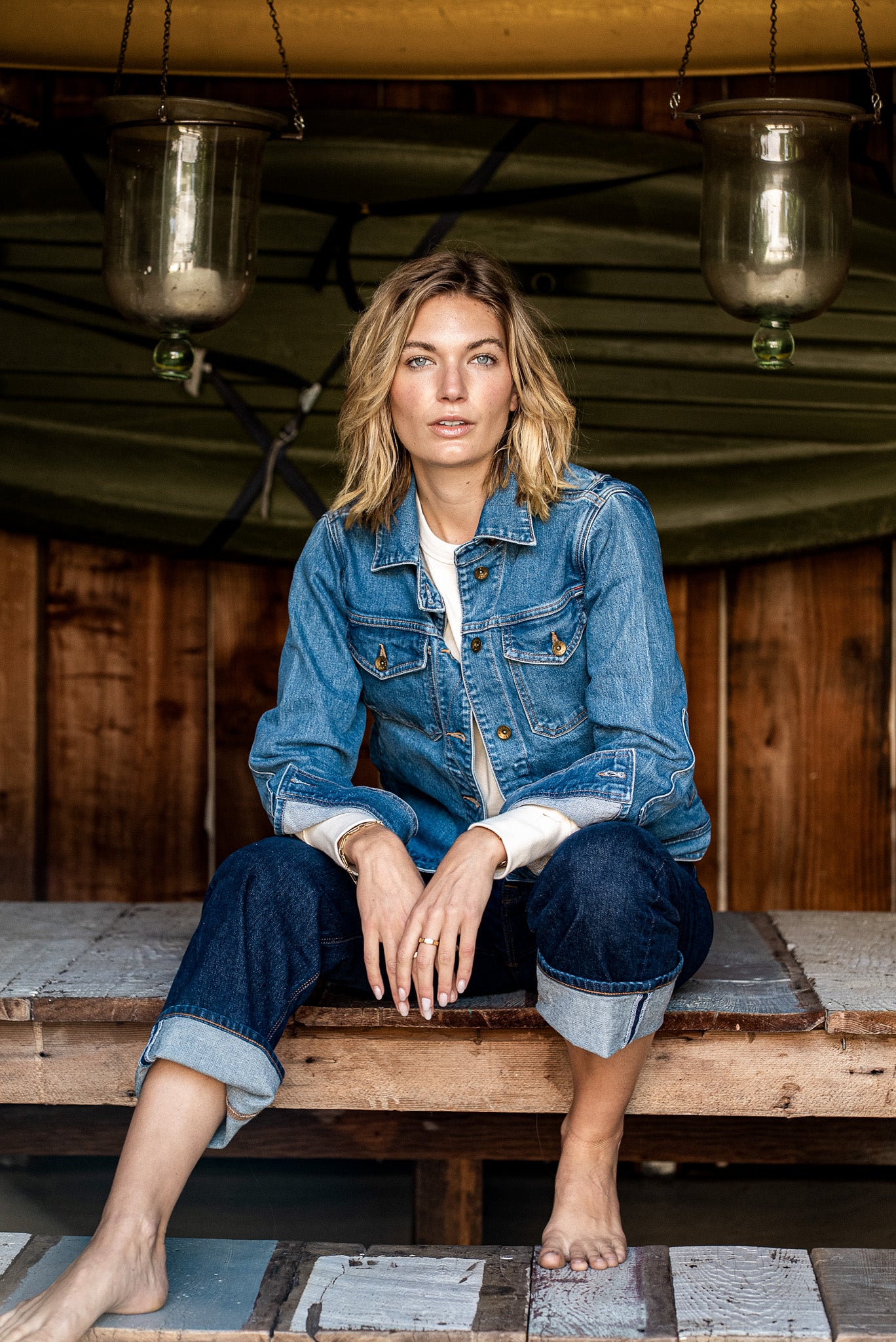 Woman in a broken twill denim jacket and cuffed jeans sitting on a wooden bench under a canoe.