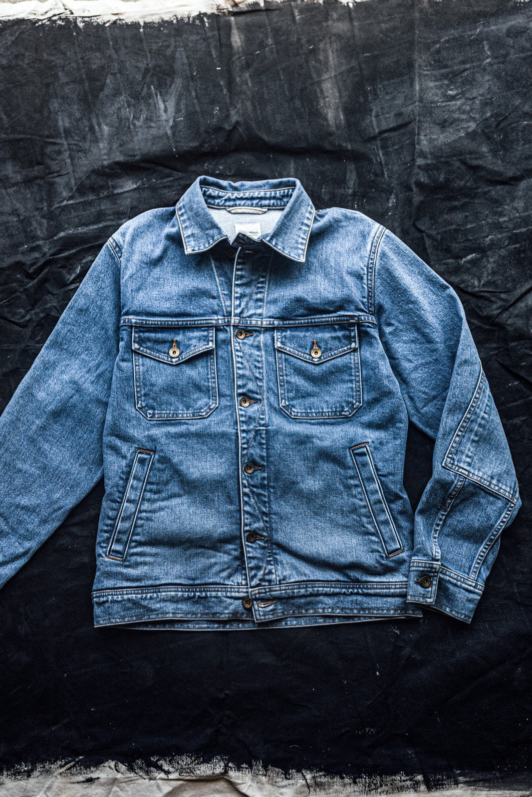 Light-wash broken twill denim jacket with chest flap pockets and brass buttons, laid flat on dark canvas.