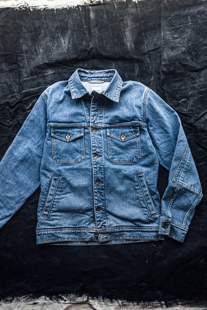 Light-wash broken twill denim jacket with chest flap pockets and brass buttons, laid flat on dark canvas.
