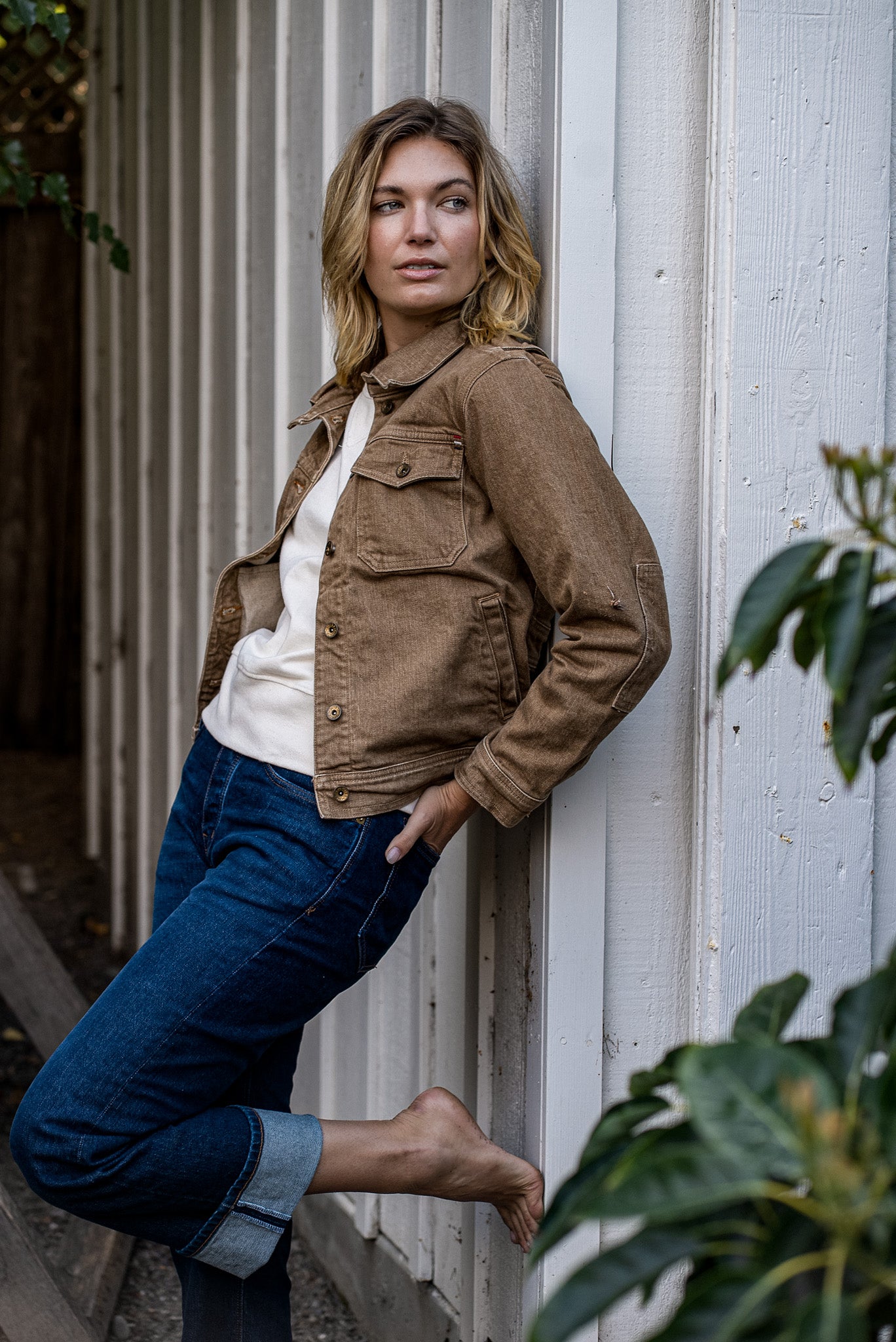 Woman leaning against a white wood wall wearing the wheat broken twill denim Artcher Jacket with cuffed blue jeans.