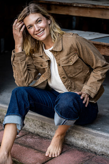 Woman in a wheat broken twill denim jacket and cuffed selvedge jeans seated on a brick step.