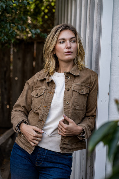 Woman wearing a wheat-colored broken twill denim jacket over a cream tee and blue jeans, leaning against a white wooden door.