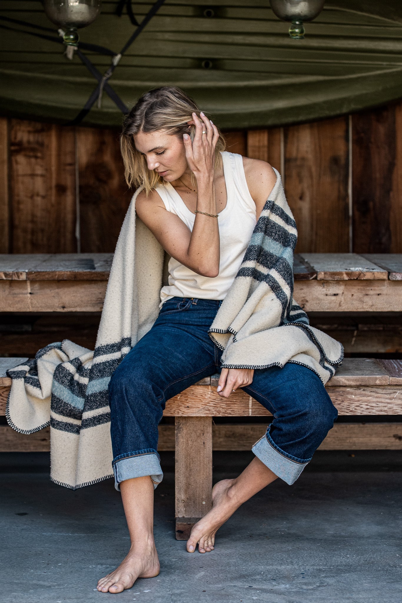 Model in natural cotton Reyes tank and cuffed denim jeans, seated on a wooden bench with a striped wool blanket.