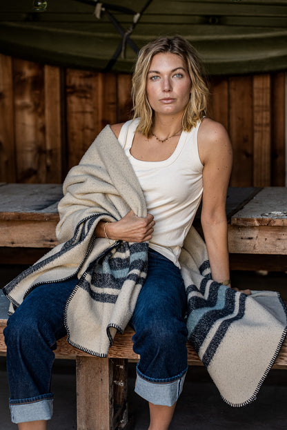 Woman in natural cotton Reyes tank and cuffed denim jeans, wrapped in a wool blanket on a wooden bench.