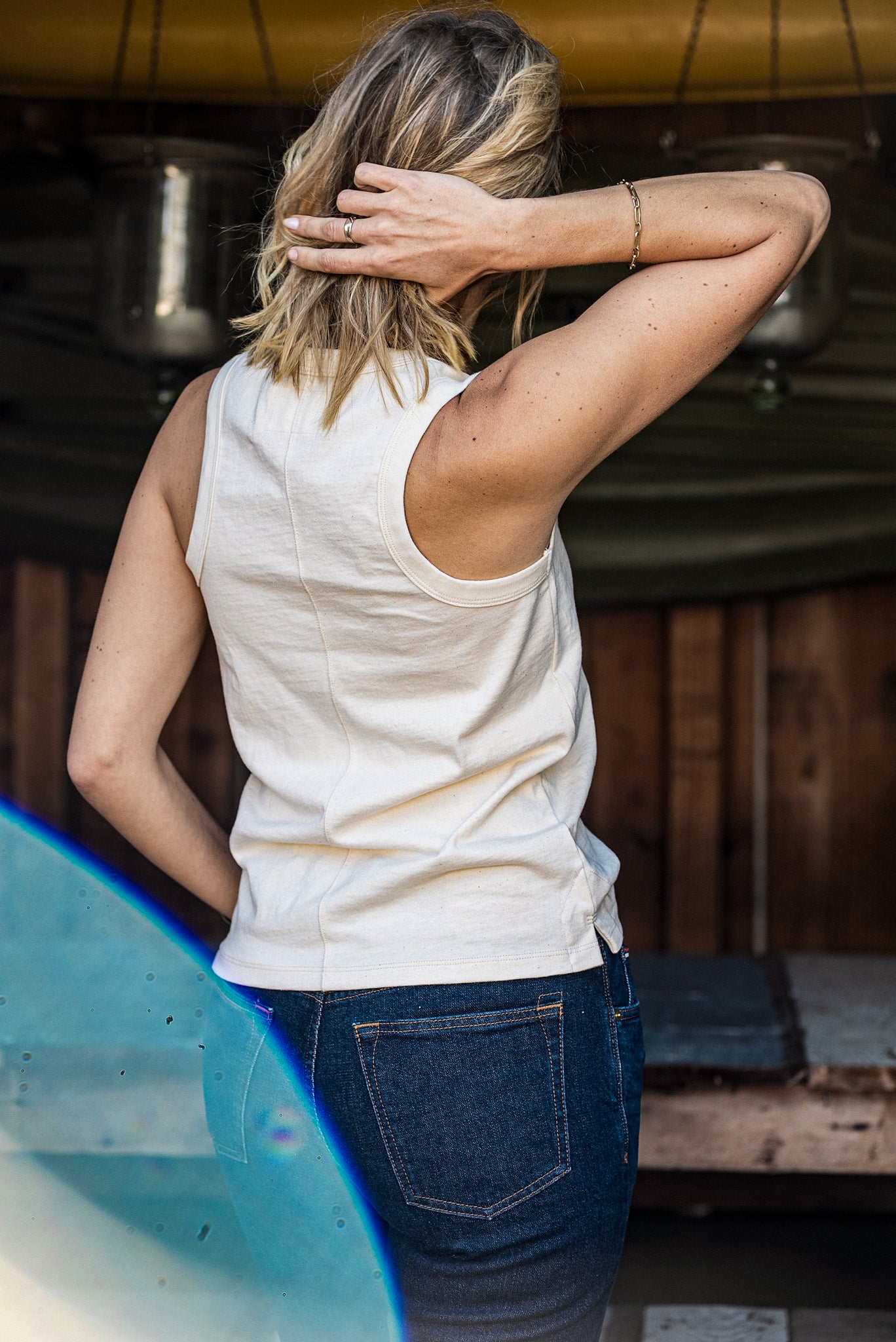 Back view of the natural cotton Reyes tank paired with dark denim jeans and a surfboard.